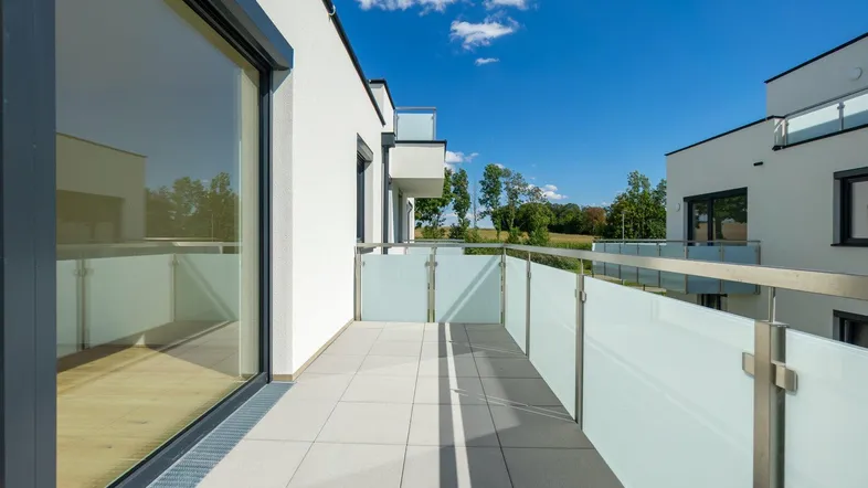 Heller Balkon mit moderner Glasbrüstung, Fliesenboden und Blick ins Grüne.