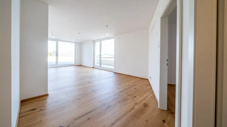 Helles, leeres Zimmer mit Holzboden und großen Fenstern, die viel Tageslicht hereinlassen.