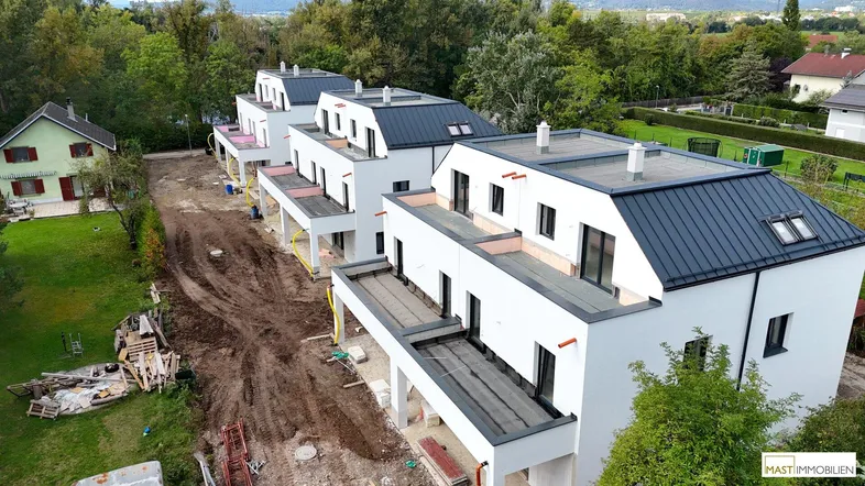 Mehrere moderne Reihenhäuser im Bau mit weißer Fassade und dunklen Dächern in grüner Umgebung.