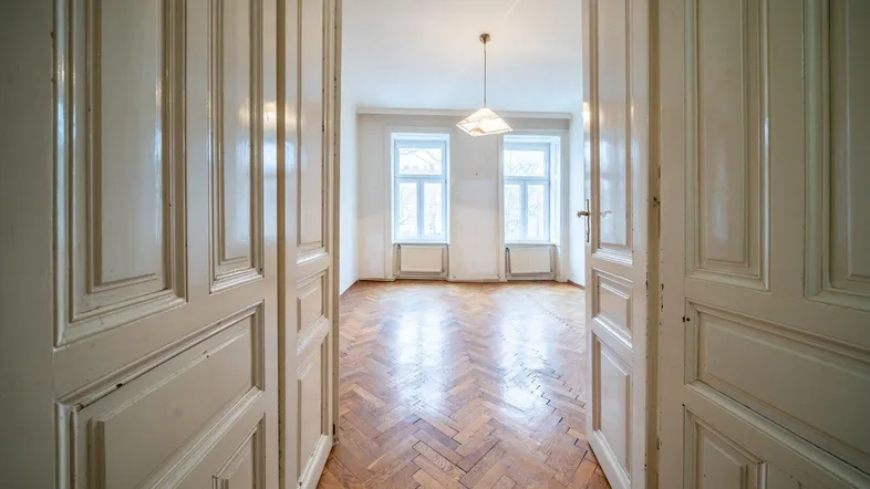 Geräumiges, leeres Zimmer mit Fischgrätparkett und hohen Doppeltüren, bereit zur Renovierung.