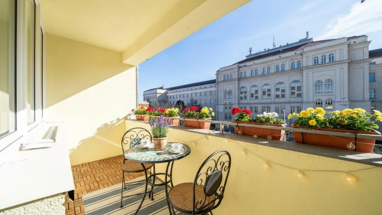 Gemütlicher Balkon mit Holzboden, Blumenkästen und Blick auf ein historisches Gebäude.