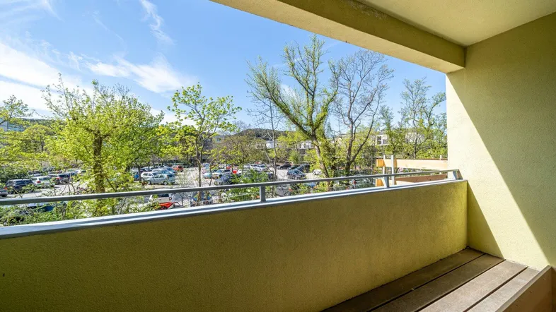 Heller Balkon mit Holzboden und Blick auf den Parkplatz und grüne Bäume.