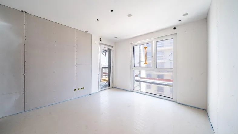 Heller Innenraum im Rohbau mit großen Fenstern und Zugang zu einem potenziellen Balkon.