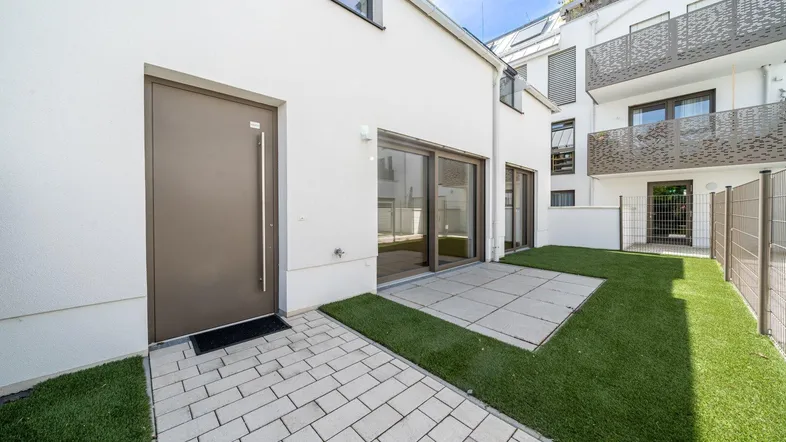 Eingangsbereich eines modernen Hauses mit kleiner gepflasterter Terrasse und Kunstrasenfläche.