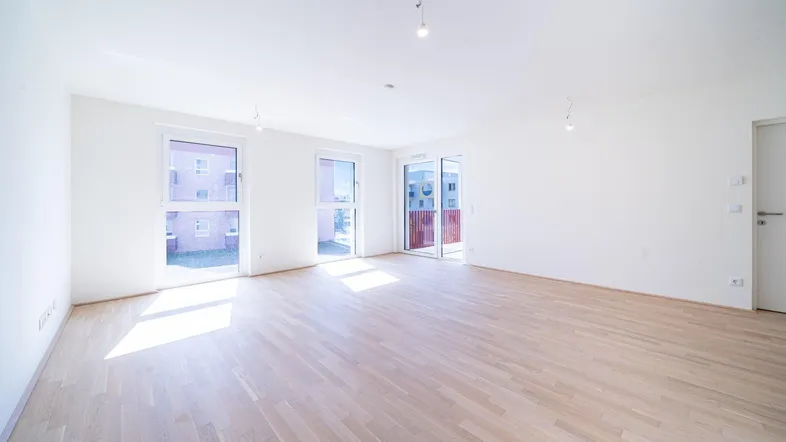 Helles, leeres Wohnzimmer mit Parkettboden und Zugang zu einem Balkon.