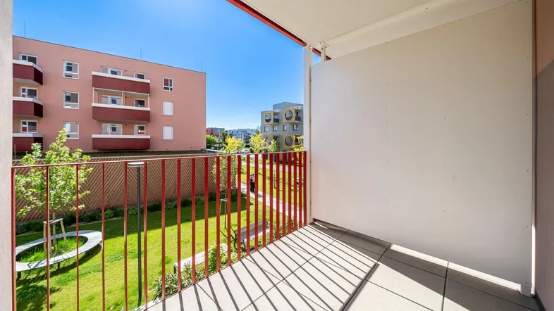 Heller Balkon mit Fliesenboden und rotem Geländer, bietet Blick auf den Gemeinschaftsgarten und umliegende Gebäude.