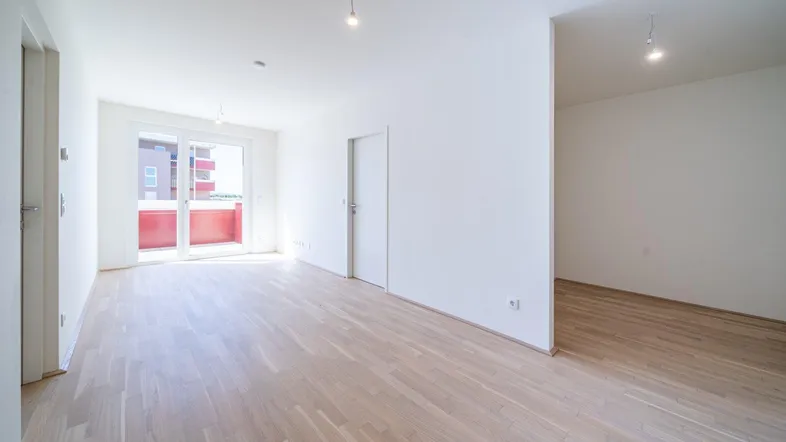Helles, leeres Zimmer mit Parkettboden und Zugang zu einem roten Balkon, ideal für Wohn- oder Schlafbereich.