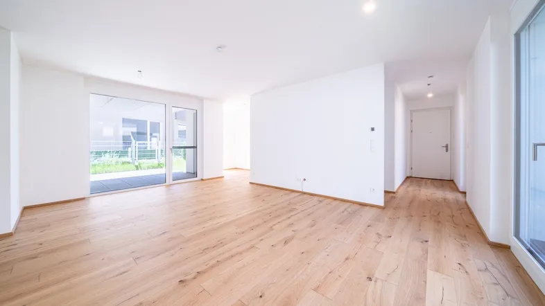 Helles, leeres Wohnzimmer mit Holzboden und Zugang zu einer Terrasse oder Garten.