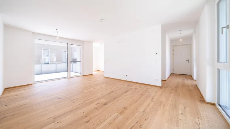 Helles, leeres Zimmer mit Holzboden und Zugang zu einem Balkon, ideal für einen Wohnbereich.