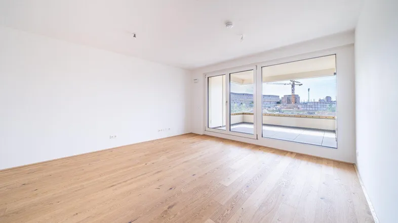 Helles, leeres Zimmer mit Holzboden und Zugang zu einem Balkon, ideal als Wohn- oder Schlafzimmer.