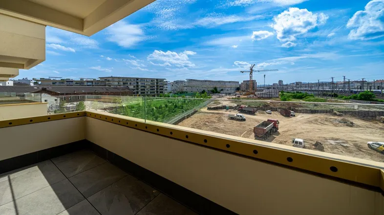 Geräumiger Balkon mit Fliesenboden und Glasgeländer, bietet einen weiten Blick über die Stadt und eine Baustelle.