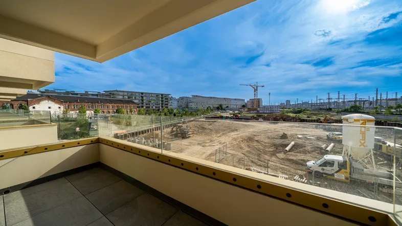 Heller Balkon mit Fliesenboden und Glasgeländer, bietet einen weiten Blick über eine Baustelle.