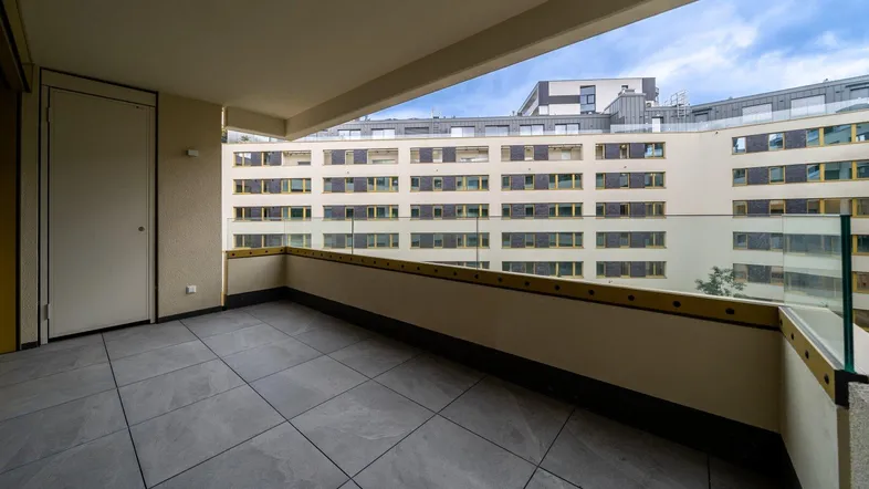 Geräumiger Balkon mit Fliesenboden und Glasgeländer, bietet Blick auf umliegende Gebäude.