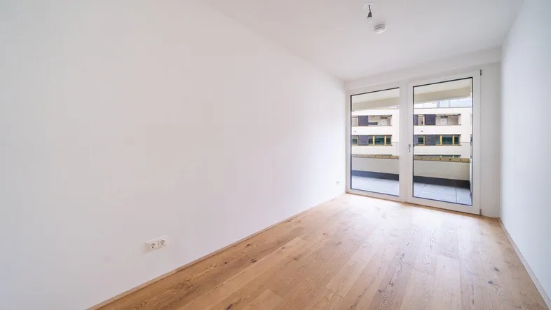 Helles, leeres Zimmer mit Holzboden und Zugang zu einem Balkon, ideal für Wohn- oder Schlafbereich.