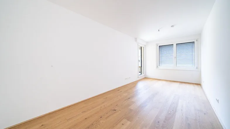 Helles, leeres Zimmer mit Parkettboden und Zugang zu einem sonnigen Außenbereich.