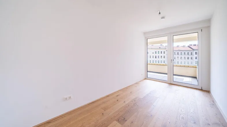 Helles Zimmer mit Holzboden und Zugang zu einem Balkon, bietet einen Blick auf die umliegenden Gebäude.