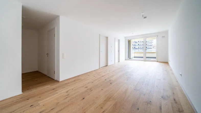 Heller, leerer Raum mit modernem Parkettboden und Zugang zu einem Balkon in einem Neubau.