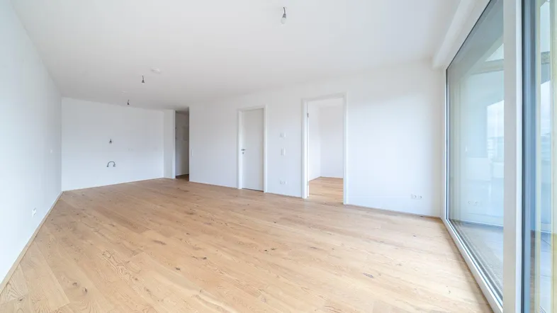 Helles, leeres Wohnzimmer mit Parkettboden und Zugang zu einem großen Balkon.
