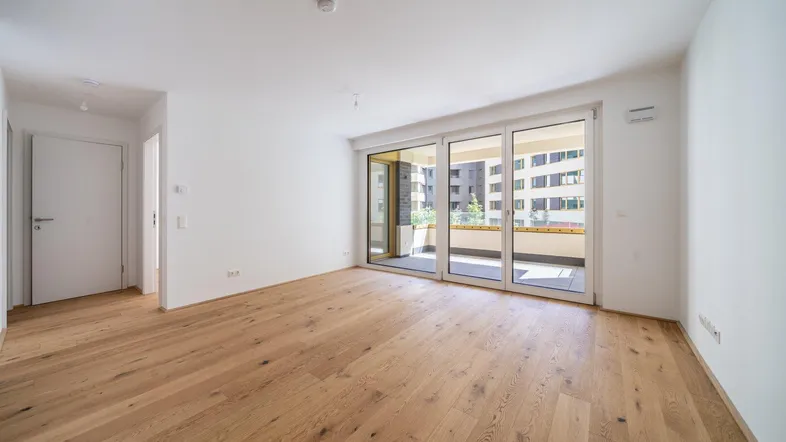 Helles, leeres Zimmer mit Holzboden und Zugang zu einem Balkon, ideal als Wohnbereich.