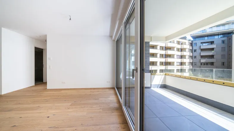 Heller, leerer Wohnraum mit Holzboden und Zugang zu einem gefliesten Balkon mit Blick auf andere Gebäude.