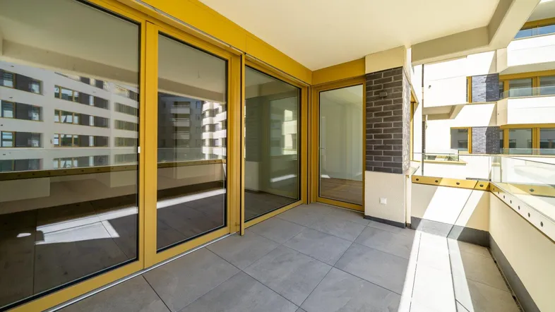Heller Balkon mit Fliesenboden und Glasgeländer, bietet einen sonnigen Außenbereich mit Blick auf umliegende Gebäude.