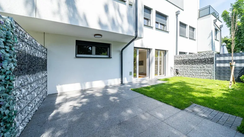 Moderne Hausfassade mit gepflasterter Terrasse, Rasenfläche und Gabionenwänden im Außenbereich.
