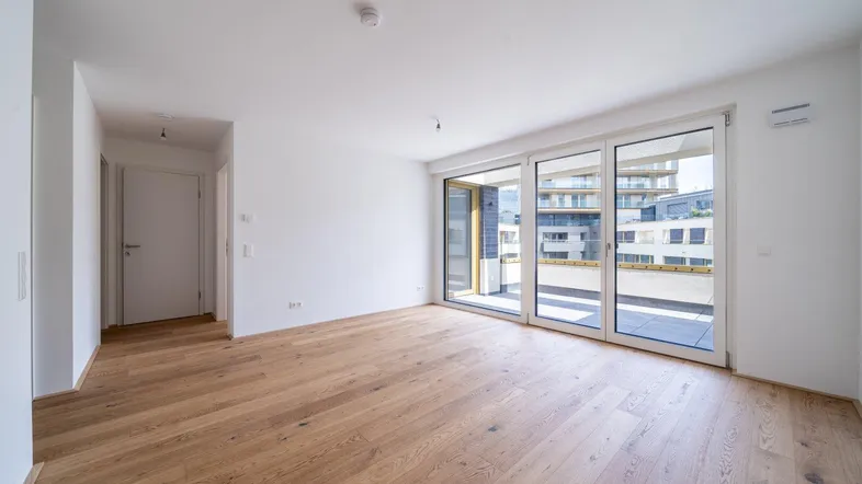Helles, unmöbliertes Wohnzimmer mit Holzboden und großen Fenstern, die auf einen Balkon führen.