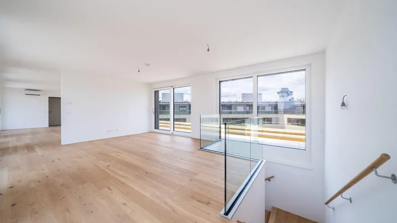 Heller, leerer Wohnbereich mit Holzboden, großen Fenstern und Zugang zu einer Treppe.