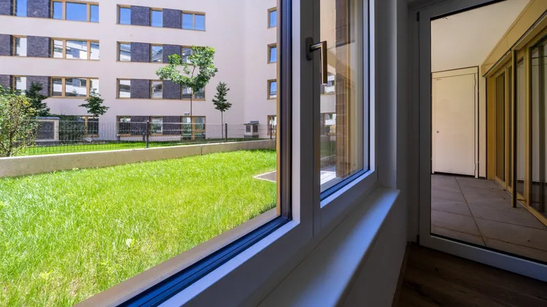 Blick aus einem Innenraum durch ein großes Fenster auf einen grünen Innenhof und moderne Wohngebäude.