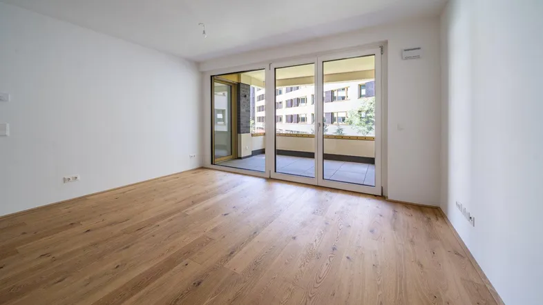 Helles Zimmer mit neuem Parkettboden und Zugang zu einem Balkon, mit Blick auf umliegende Gebäude.