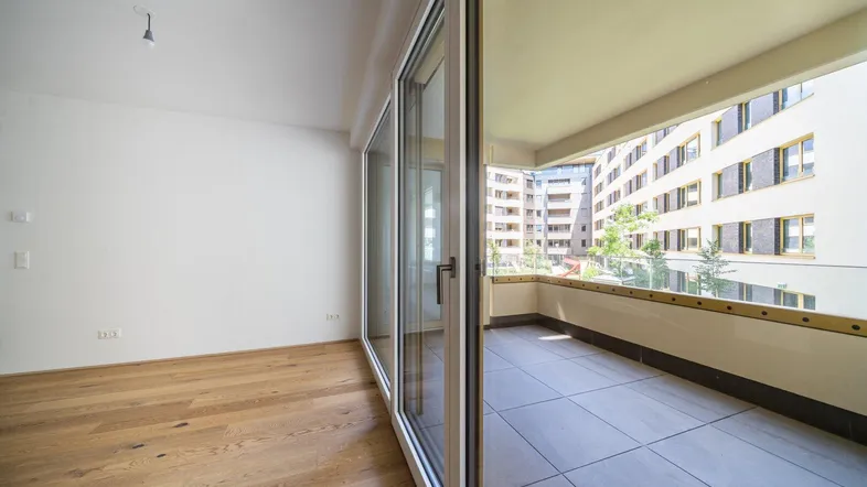 Heller Raum mit Holzboden und großen Schiebetüren, die zu einem gefliesten Balkon führen.