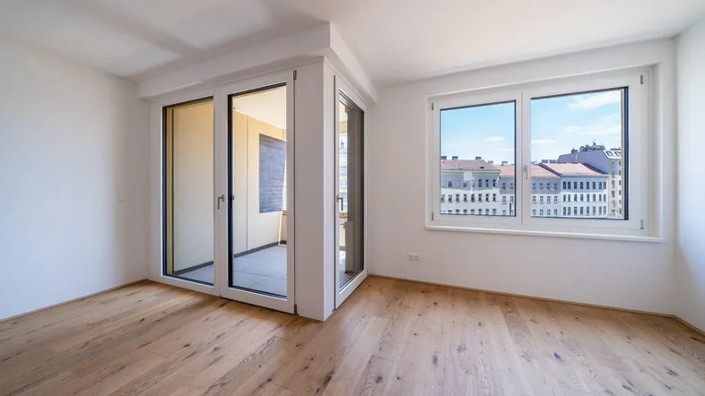 Helles, leeres Zimmer mit Parkettboden und Zugang zu einem Balkon, bietet einen Stadtblick.