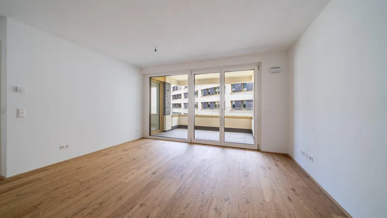 Helles, leeres Zimmer mit Holzboden und großen Fenstern, die auf einen Balkon führen.