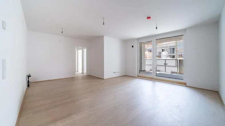 Helles, leeres Wohnzimmer mit modernem Holzfußboden und Zugang zu einem Balkon.