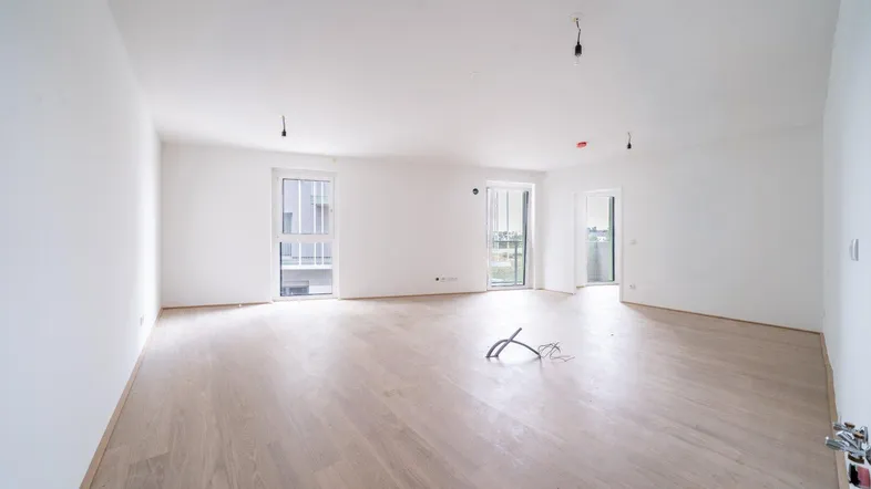 Helles, leeres Wohnzimmer mit hellem Holzfußboden und mehreren Fenstern, die viel Tageslicht hereinlassen.