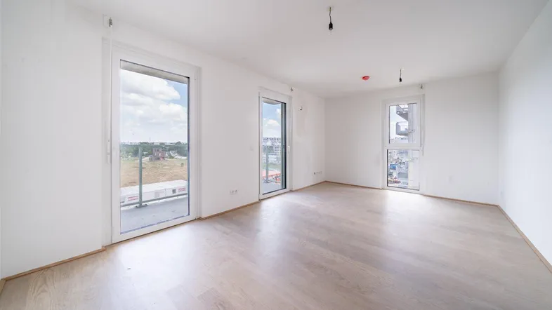 Helles, leeres Zimmer mit hellem Holzboden und großen Fenstern, die viel Tageslicht hereinlassen.