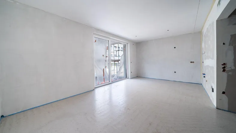 Unfertiger Innenraum mit Rohbauwänden und Zugang zu einem Balkon oder einer Terrasse durch große Fenster.