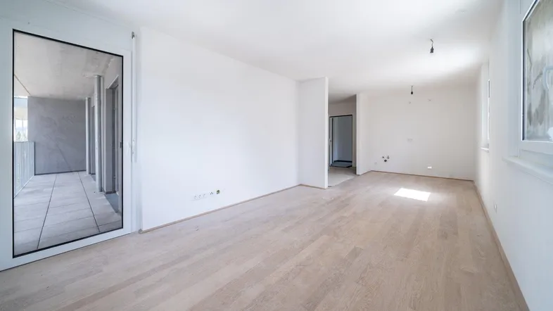 Helles, leeres Wohnzimmer mit hellem Holzboden und Zugang zu einem Balkon, in neuwertigem Zustand.