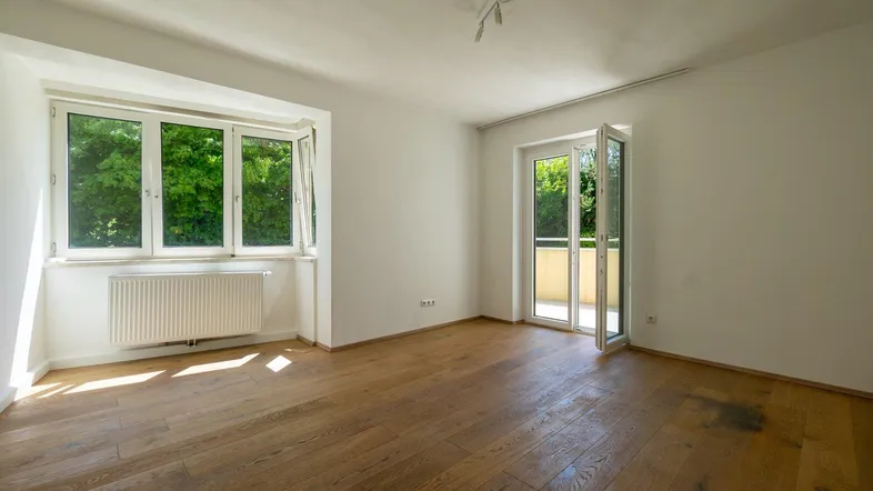 Helles Zimmer mit Holzboden, großem Fenster und Zugang zu einem Balkon mit grüner Aussicht.