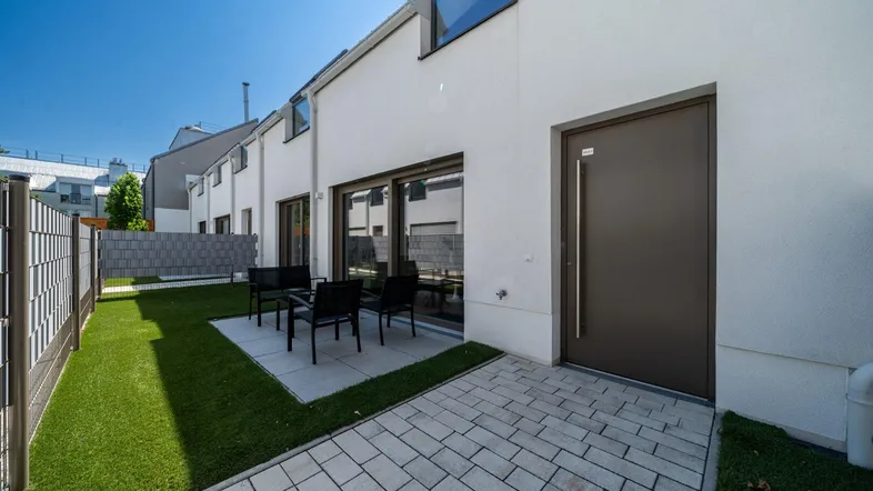Moderne Reihenhausfassade mit kleiner Terrasse, Kunstrasenfläche und Sitzgelegenheiten im Freien.