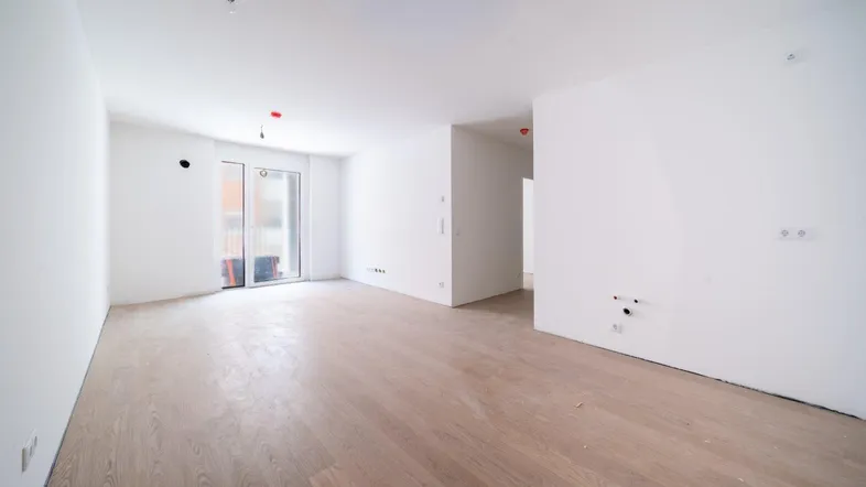 Helles, leeres Wohnzimmer mit neuem Parkettboden und Zugang zu einem Balkon oder einer Terrasse.