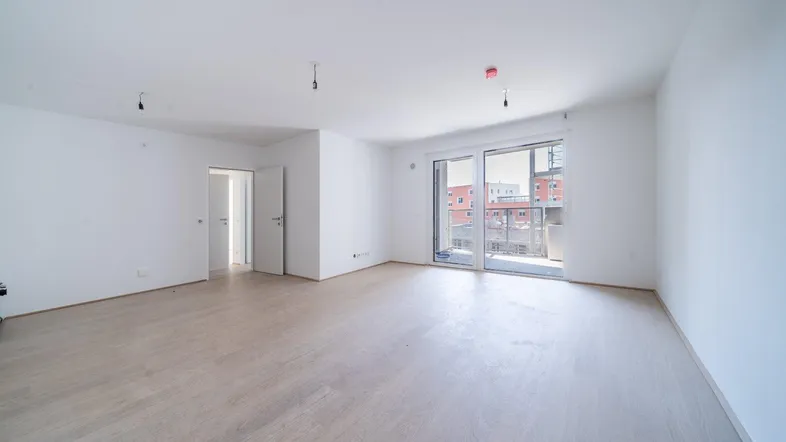 Helles, leeres Wohnzimmer mit hellem Parkettboden und Zugang zu einem Balkon.