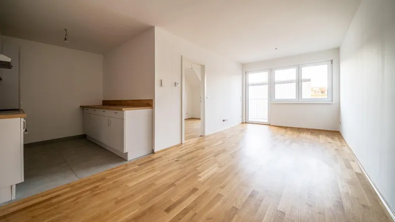 Heller offener Wohnbereich mit Parkettboden und Zugang zu einem Balkon, angrenzend an eine Küchenzeile.