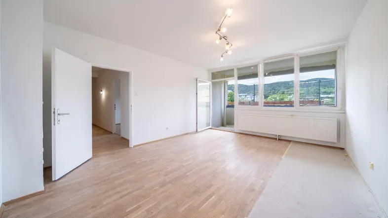 Helles Wohnzimmer mit Parkettboden, großen Fenstern und Zugang zu einem Balkon mit Grünblick.