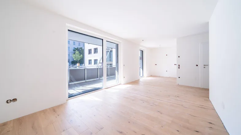 Helles, leeres Zimmer mit Holzboden und Zugang zu einem Balkon, bereit für die Einrichtung.