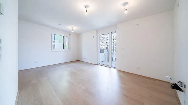 Helles, leeres Wohnzimmer mit hellem Parkettboden und Zugang zu einem Balkon in einem Neubau.