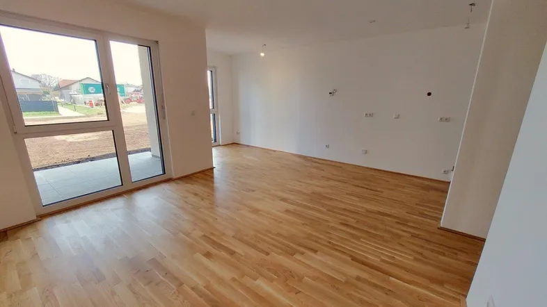 Helles, unmöbliertes Wohnzimmer mit großen Fenstern, Holzboden und Zugang zum Außenbereich.