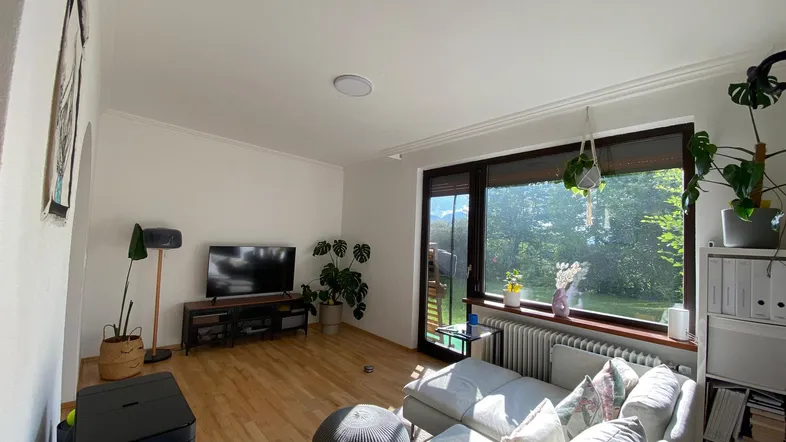 Helles Wohnzimmer mit Holzboden, großem Fenster und Blick ins Grüne, ausgestattet mit Sofa und TV.