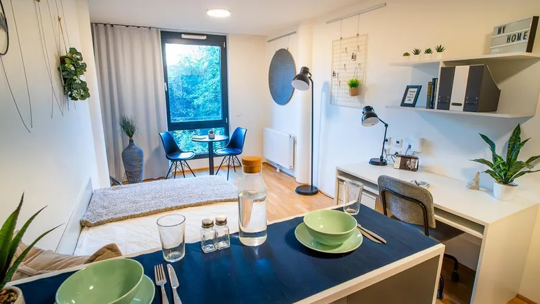Helles, modernes Schlafzimmer mit integriertem Arbeitsbereich, großem Fenster und kleinem Essbereich.