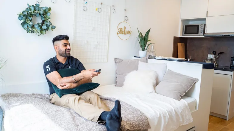 Junger Mann entspannt auf dem Bett in einem modernen Studio-Apartment mit integrierter Küchenzeile.